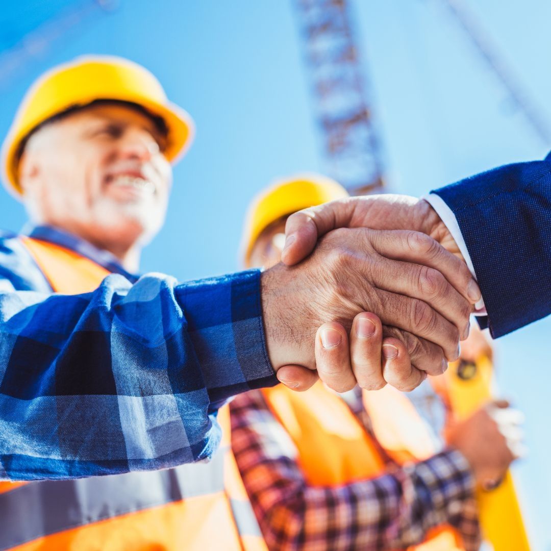Zwei Personen mit Schutzhelmen und Warnwesten reichen sich auf einer Baustelle die Hand